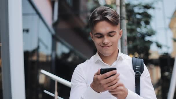 Glad ung affärsman eller turist med en ryggsäck som använder telefonen stående nära modern kontorsbyggnad. Koncept: teknik, telefoni, affärsresor, business — Stockvideo