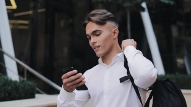 Mutlu genç işadamı veya turist modern Ofis Binası yakınındaki yürümek telefon kullanarak bir sırt çantası ile. Konsept: teknoloji, telefon, iş gezileri, iş