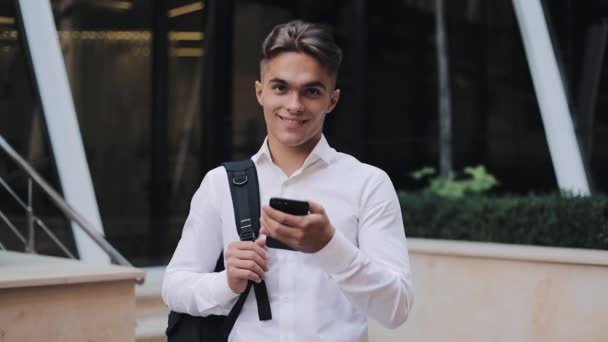 Guapo joven empresario o turista sosteniendo un teléfono en las manos, mirando a la cámara y sonriendo. Concepto: nuevos negocios, viajes por el mundo, comunicación, contactos, comunicaciones, negocios — Vídeos de Stock