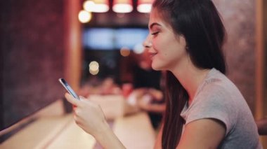 Genç kadın otururken bar, lemanade içme ve neon bar işareti yanındaki akıllı telefonu kullanarak. Kadının onun smartphone üzerinde arkadaşlarınızla iletişim sırasında gece hayatı