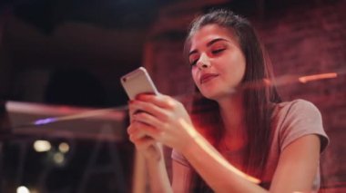 Genç çekici güzel gülümseme ile Smartphone kullanarak akşam saatlerinde kadındır. Seksi kız, bir bar veya restoran neon tabela yakın oturan
