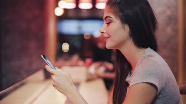 Молодая женщина сидит в баре, пьет лиманад и пользуется смартфоном рядом с вывеской неонового бара. Женщина наслаждается ночной жизнью, общаясь с друзьями на смартфоне — стоковое видео