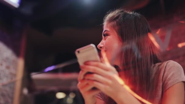 Mujer atractiva joven con hermosa sonrisa está utilizando el teléfono inteligente en la noche. Chica sexy sentada en un bar o restaurante cerca de la señalización de neón — Vídeo de stock