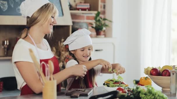 Mamá y su hijita cocinan en la cocina: hacen una ensalada de jardín fresco, y la madre enseña a la niña. — Vídeo de stock
