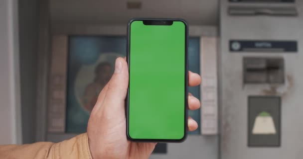 Mani maschili che tengono smartphone nero con schermo verde sullo sfondo ATM. Grande schermo. Chiave cromatica. Da vicino. — Video Stock