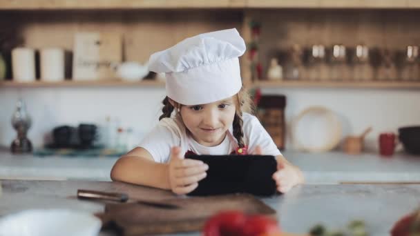 Linda niña utilizando el teléfono inteligente durante el desayuno sentado en la cocina. Ella está vestida con un delantal mirando la pantalla del teléfono inteligente — Vídeos de Stock