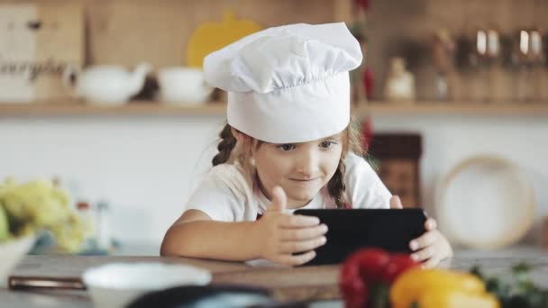 Roztomilá holčička pomocí smartphone během snídaně sedí na kuchyni. Je oblečen v zástěře při pohledu na obrazovku smartphone