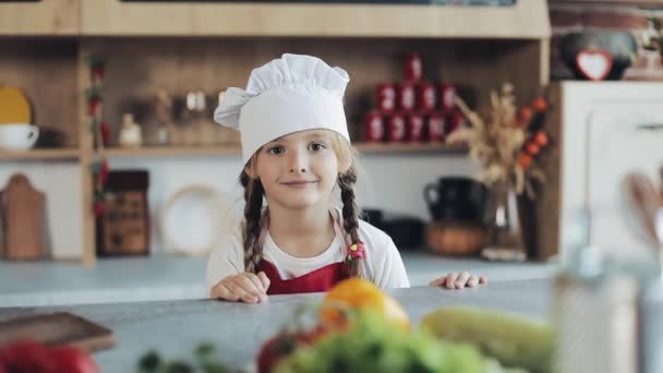 Porträtt av en söt liten flicka i köket klädd som en professionell kock tittar in i kameran och leende. Begreppet: näringslära, Matlagning skola, utbildning — Stockvideo