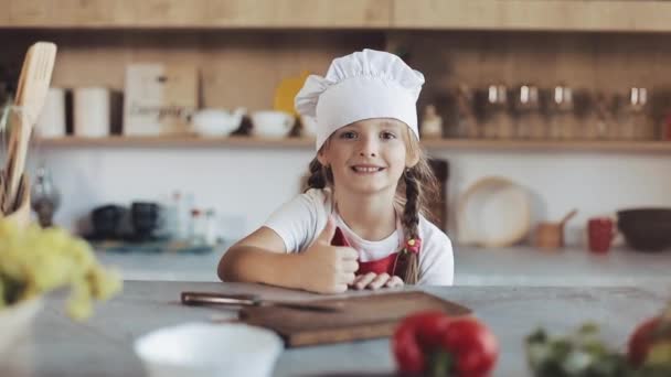 Ritratto di una graziosa bambina in cucina vestita da cuoca professionista che guarda nella macchina fotografica e sorride. Mostra un pollice in su. Concetto di: nutrizione, scuola di cucina, educazione — Video Stock