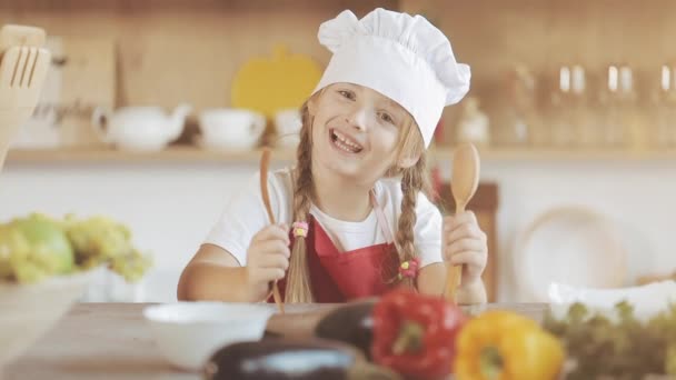 Ritratto di una graziosa bambina in cucina vestita da cuoca professionista che guarda nella macchina fotografica e sorride. Lei tiene in mano una forchetta e un cucchiaio e si diverte . — Video Stock