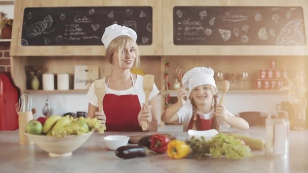 Mom and little daughter in the kitchen dressed as a professional cooks looking into the camera and smiling. They holds a fork and a spoon in her hands and have fun — Stock Video