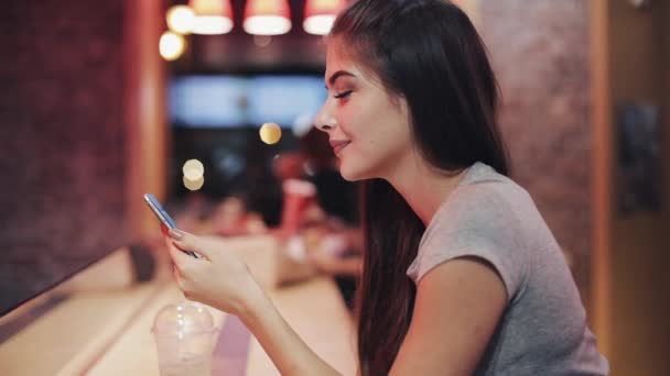 Jovem mulher atraente com sorriso bonito está usando Smartphone na noite e olhando para a câmera. Menina sexy sentado em um bar ou restaurante perto de sinalização de néon — Vídeo de Stock