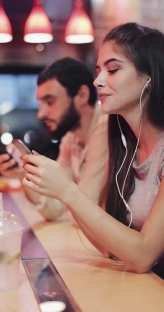 Vídeo vertical. Hermosa joven sentada detrás de un bar, usando un teléfono inteligente, escuchando música, sonriendo, bebiendo un cóctel. Ella se sienta al lado de un joven . — Vídeos de Stock