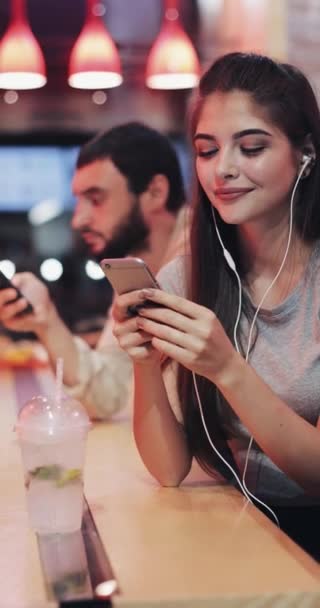 Κάθετη βίντεο. Όμορφη νεαρή γυναίκα κάθεται πίσω από ένα μπαρ, χρησιμοποιώντας ένα smartphone, ακούγοντας μουσική, χαμογελώντας, πίνοντας ένα κοκτέιλ. Κάθεται δίπλα σε έναν νεαρό άνδρα. — Αρχείο Βίντεο