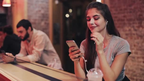 Aantrekkelijke jonge vrouw jonge vrouw met hoofdtelefoon in haar oren luisteren naar muziek en het gebruik van de smartphone. Wazig nacht. Ze zit naast een jonge man — Stockvideo