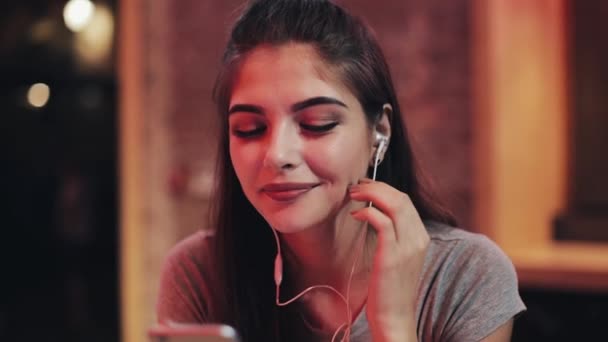 Jovem mulher atraente com fones de ouvido nos ouvidos ouvindo música e usando smartphone. Noite desfocada. Ela se senta ao lado de um jovem — Vídeo de Stock