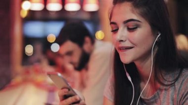 Çekici genç kadın müzik ve smartphone kullanarak kulaklarını kulaklıklarla. Bulanık gece. O genç bir adamın yanına oturur
