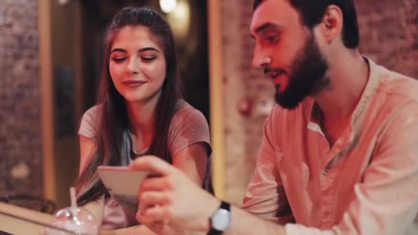 Glückliches junges Paar mit Smartphone plaudernd und Getränk an der Bar sitzend. Freunde, die gemeinsam ihr Handy-Smartphone benutzen, lächeln und lachen bei einem Date — Stockvideo