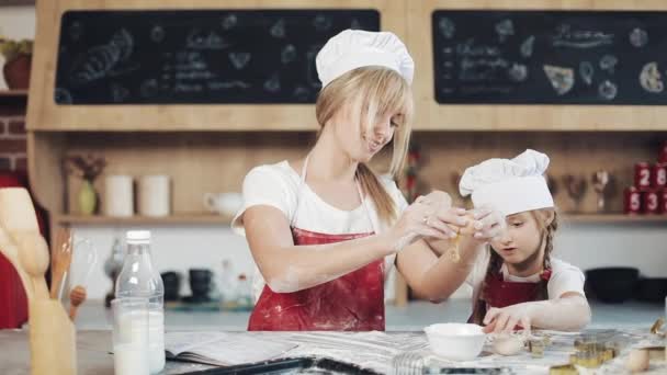 Mamá e hija con la misma ropa se divierten preparando una masa en una cocina acogedora. Están rompiendo huevos en un tazón. Cocina, Concepto de mamá e hija — Vídeos de Stock
