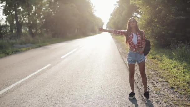 Viajero joven mujer autostop en un camino soleado. Joven chica sexy en busca de un paseo para iniciar un viaje. Hora de verano — Vídeo de stock