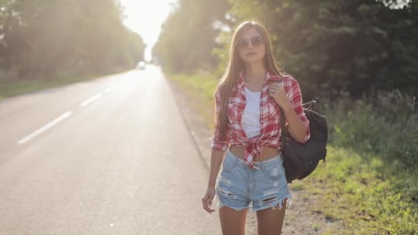 日当たりの良い道路でヒッチハイク旅行者の若い女性。乗車時間を探して、旅を開始する若いセクシーな女の子夏の時間 — ストック動画