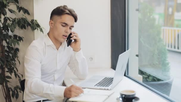 Средний снимок концентрированного молодого бизнесмена, делающего заметки в блокноте во время разговора по телефону во время кофе-брейка — стоковое видео