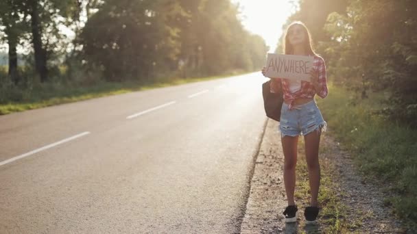 Mulher bonita jovem carona em pé na estrada segurando em qualquer lugar sinal. Hora de verão — Vídeo de Stock