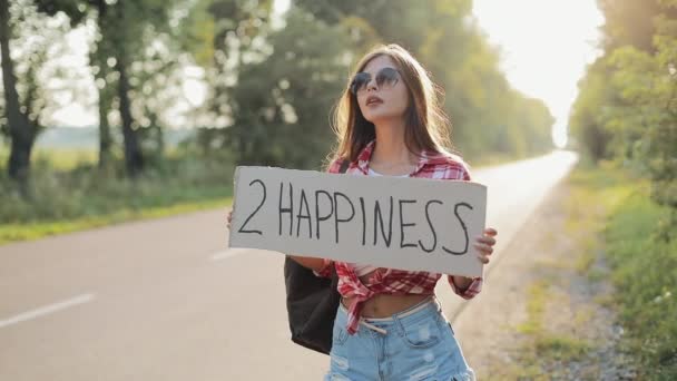 Молодая красивая женщина автостопом стоя на дороге держа 2 знак счастья. Летнее время — стоковое видео