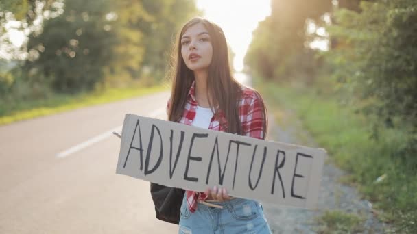 Young beautiful woman hitchhiking standing on the road holding adventure sign. Summer time. — Stock Video