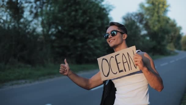 Jovem homem atraente carona em pé na estrada segurando sinal do oceano. Hora de verão — Vídeo de Stock