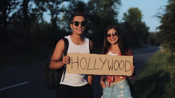 Um jovem casal está a pedir boleia de pé na estrada. Um homem e uma mulher param o carro na auto-estrada com um sinal de Hollywood. A pedir boleia. Viajante. Serviços de transporte — Vídeo de Stock