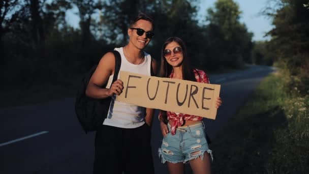 Una joven pareja está haciendo autostop de pie en la carretera. Un hombre y una mujer detienen el coche en la carretera con una señal de Futuro. Hacer autostop. Viajero. Transporte. — Vídeos de Stock