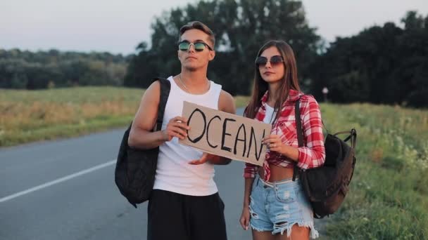 Um jovem casal está a pedir boleia de pé na estrada. Um homem e uma mulher param o carro na auto-estrada com um sinal Ocean. A pedir boleia. Viajante. Serviços de transporte — Vídeo de Stock