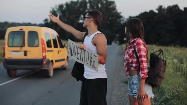 Um jovem casal está a pedir boleia de pé na estrada. Um homem e uma mulher param o carro na estrada com um sinal Adrenture. A pedir boleia. Viajante. Serviços de transporte — Vídeo de Stock