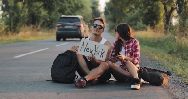 Молодая пара автостопом сидит на дороге. Мужчина и женщина останавливают машину на шоссе — стоковое видео