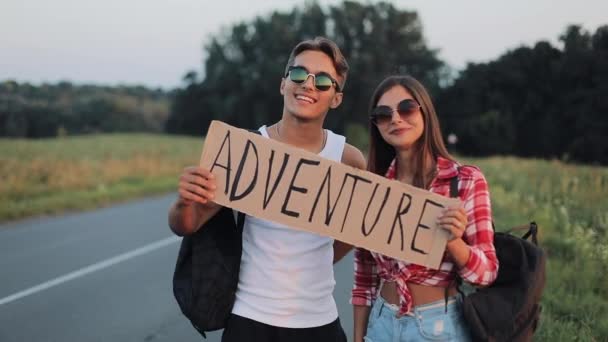 Young traveling couple in love standing on the road holding a sign Adventure. happy smiling friends are looking into the camera. Rest, trip, travel, adventure, hitchhiking concept — Stock Video