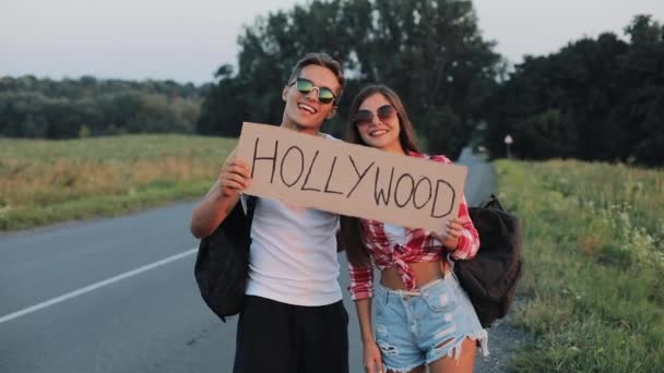 Pareja joven viajando enamorada de pie en la carretera sosteniendo un cartel de Hollywood. amigos sonrientes felices están mirando a la cámara. Descanso, viaje, aventura, concepto de autoestop — Vídeos de Stock