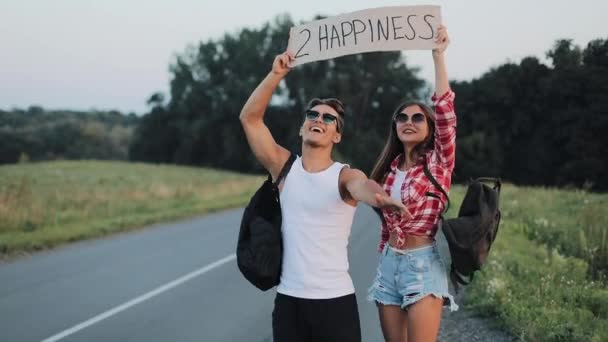Jeune couple ambulant amoureux debout sur la route tenant un signe 2 bonheur. heureux amis souriants regardent dans la caméra. Repos, voyage, aventure, concept d'auto-stop — Video