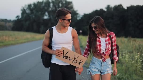 Paar im Urlaub trampt entlang der Straße und hält ein neues Schild in der Hand. Sommerzeit. Trampen, Touristen, Abenteuer — Stockvideo