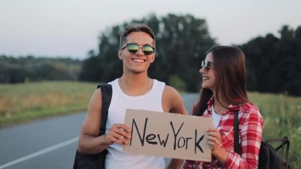 Jovem casal viajante apaixonado de pé na estrada segurando um sinal de Nova York. amigos sorridentes felizes estão olhando para a câmera. Descanso, viagem, viagem, aventura, conceito de carona — Vídeo de Stock