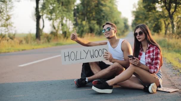 A young couple are hitchhiking sitting on the road and using smartphone. A man and a woman stop the car on the highway with a sign Hollywood — Stock Video