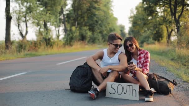 Un jeune couple fait de l'auto-stop assis sur la route et utilise un smartphone. Un homme et une femme arrêtent la voiture sur l'autoroute avec un panneau sud — Video