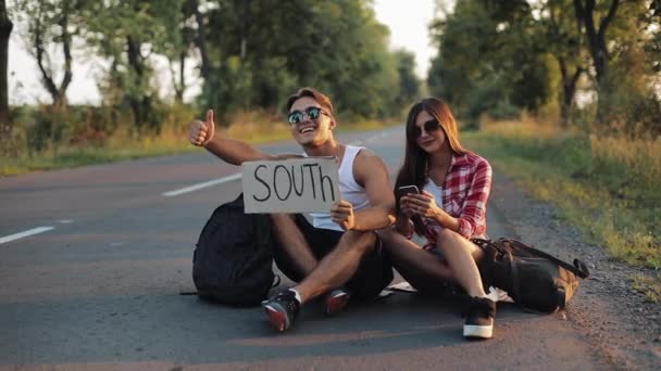 道に座っていると、スマート フォンを使用して、若いカップルがヒッチハイクします。男と女は印南で高速道路に車を止める — ストック動画
