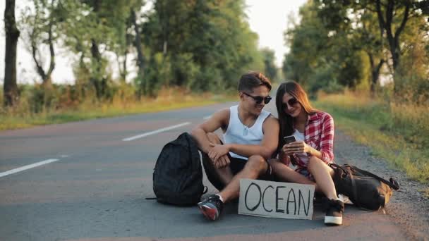 Una pareja joven está haciendo autostop sentado en la carretera y utilizando un teléfono inteligente. Un hombre y una mujer detienen el coche en la carretera con una señal Ocean — Vídeos de Stock