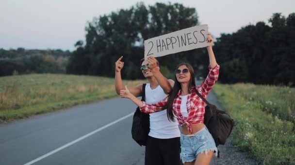 A young couple are hitchhiking standing on the road. A man and a woman stop the car on the highway with a sign 2 happiness. Hitchhiking. Traveler. Transportation — Stock Video
