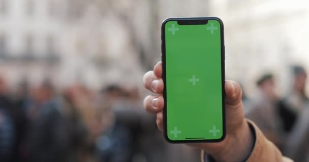 Närbild av en mans hand hållande en mobiltelefon med en vertikal grön skärm på gatorna. Militära soldater i bakgrunden — Stockvideo