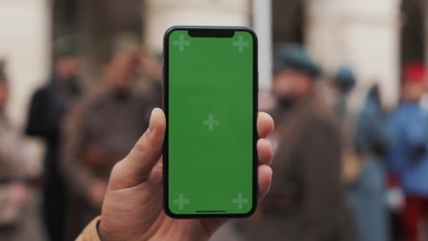 Fechar a mão de um homem segurando um telefone celular com uma tela verde vertical nas ruas. Soldados militares em segundo plano — Vídeo de Stock