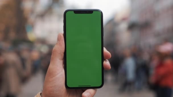 Primer plano de una mano de hombre sosteniendo un teléfono móvil con una pantalla verde vertical en las calles. Gran pantalla. Soldados militares en el fondo — Vídeos de Stock