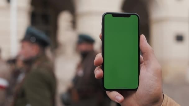 Крупним планом рука чоловіка тримає і використовує мобільний телефон з вертикальним зеленим екраном на вулицях. Військові солдати на задньому плані — стокове відео