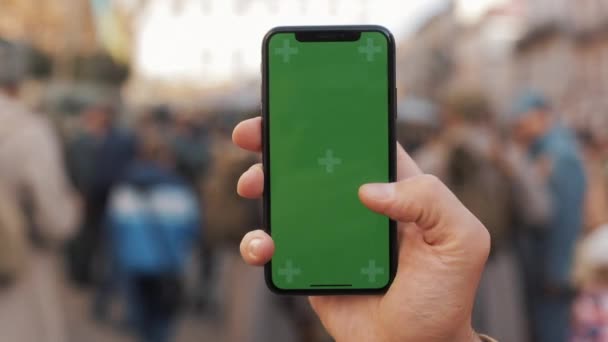 Primer plano de una mano del hombre sosteniendo y utilizando el teléfono móvil con una pantalla verde vertical en las calles. Soldados militares en el fondo — Vídeos de Stock